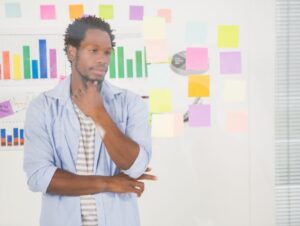 Illustration de l'article "Se libérer des conditionnements par la restructuration cognitive". Un homme pensif se tient devant une vitre en observant des notes sur des cartons colorés disposées en tableau, évoquant une énigme à résoudre avec une approche créative.