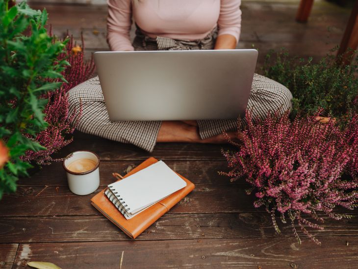 Atelier en ligne du renforcement du moi. Une femme assise en tailleur, un ordinateur sur ses jambes, a cahier et une tasse de café, dans un environnement cosy.